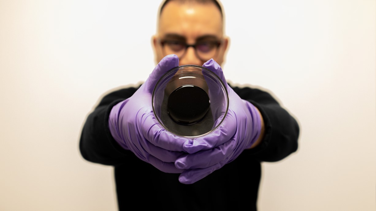 Dr Jose Norambuena-Contreras wears purple gloves as he holds a sample of self-healing bitumen in a glass dish. Credit Swansea University